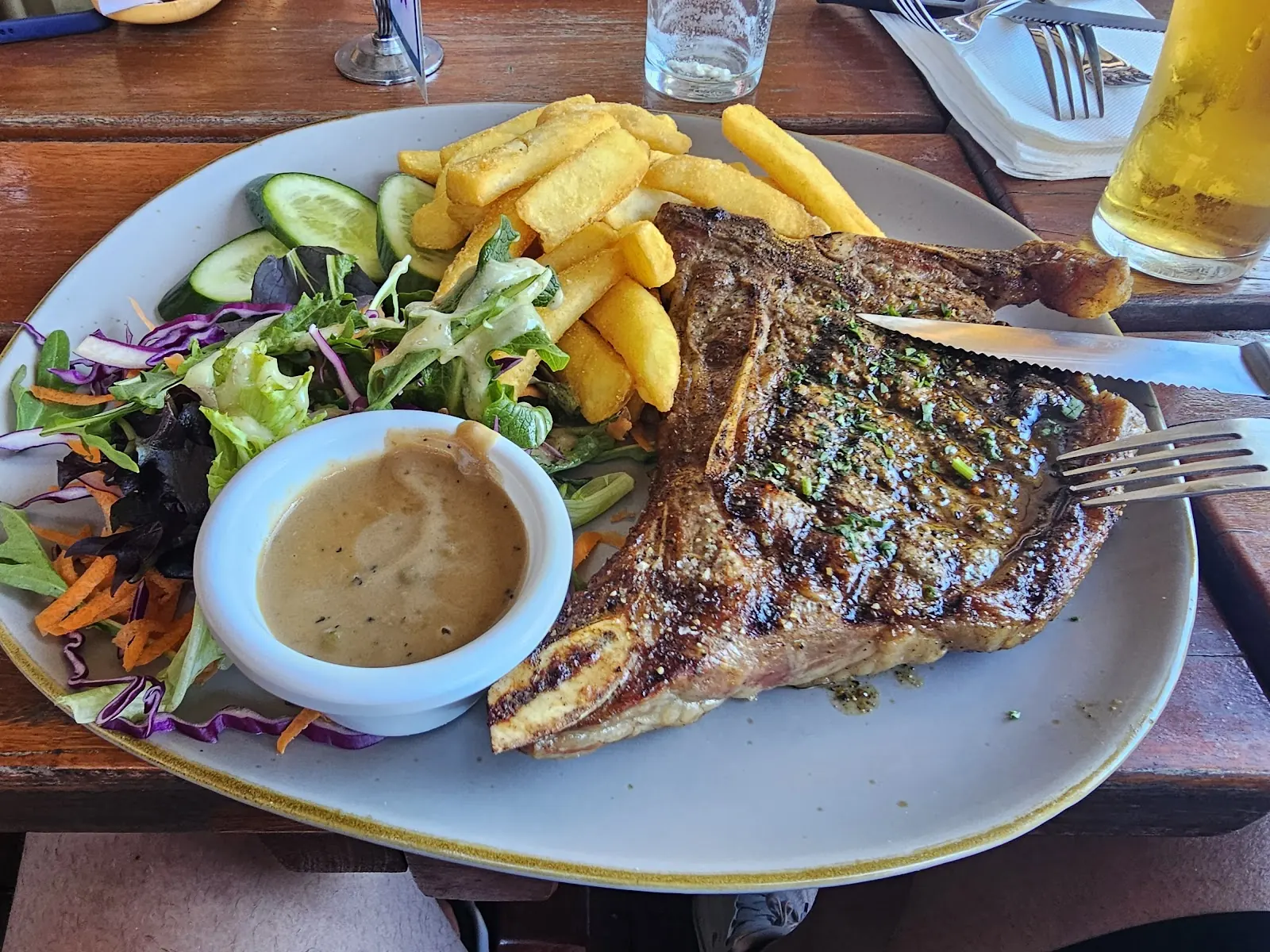 A T-bone steak with chips, salad and a pot of sauce